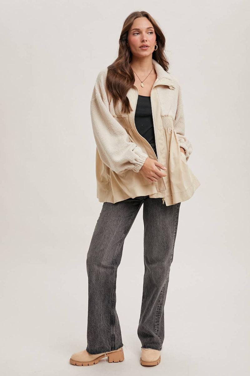 Woman wearing a beige jacket, black top, and gray pants on a white background