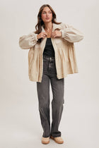 Woman wearing a beige jacket, black top, and gray jeans on a white background